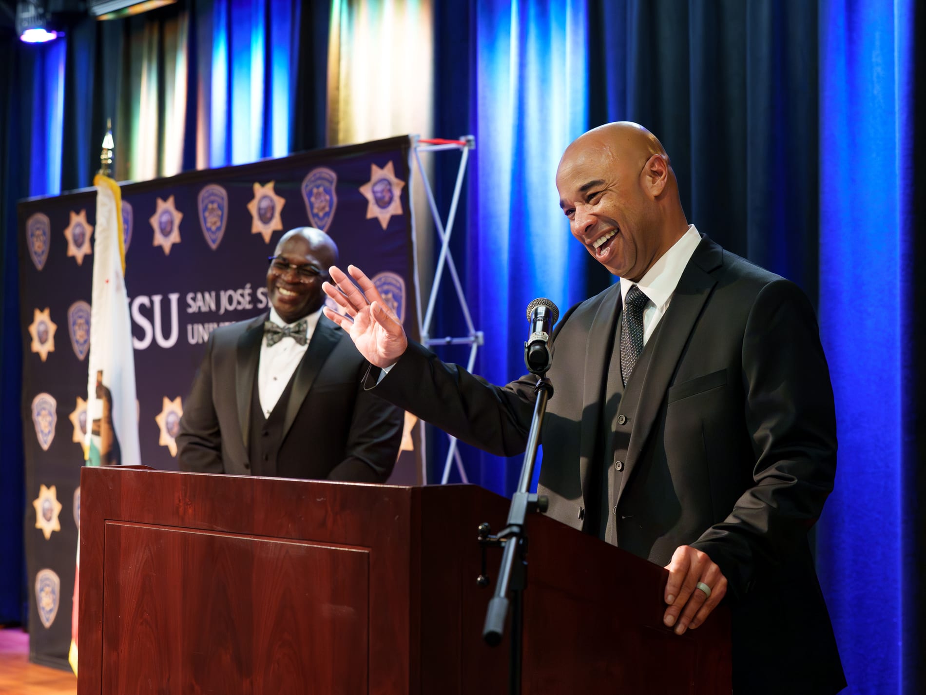 Two men share a laugh on stage, one gesturing out to the crowd, in front of an SJSU San Jose State University police badge backdrop