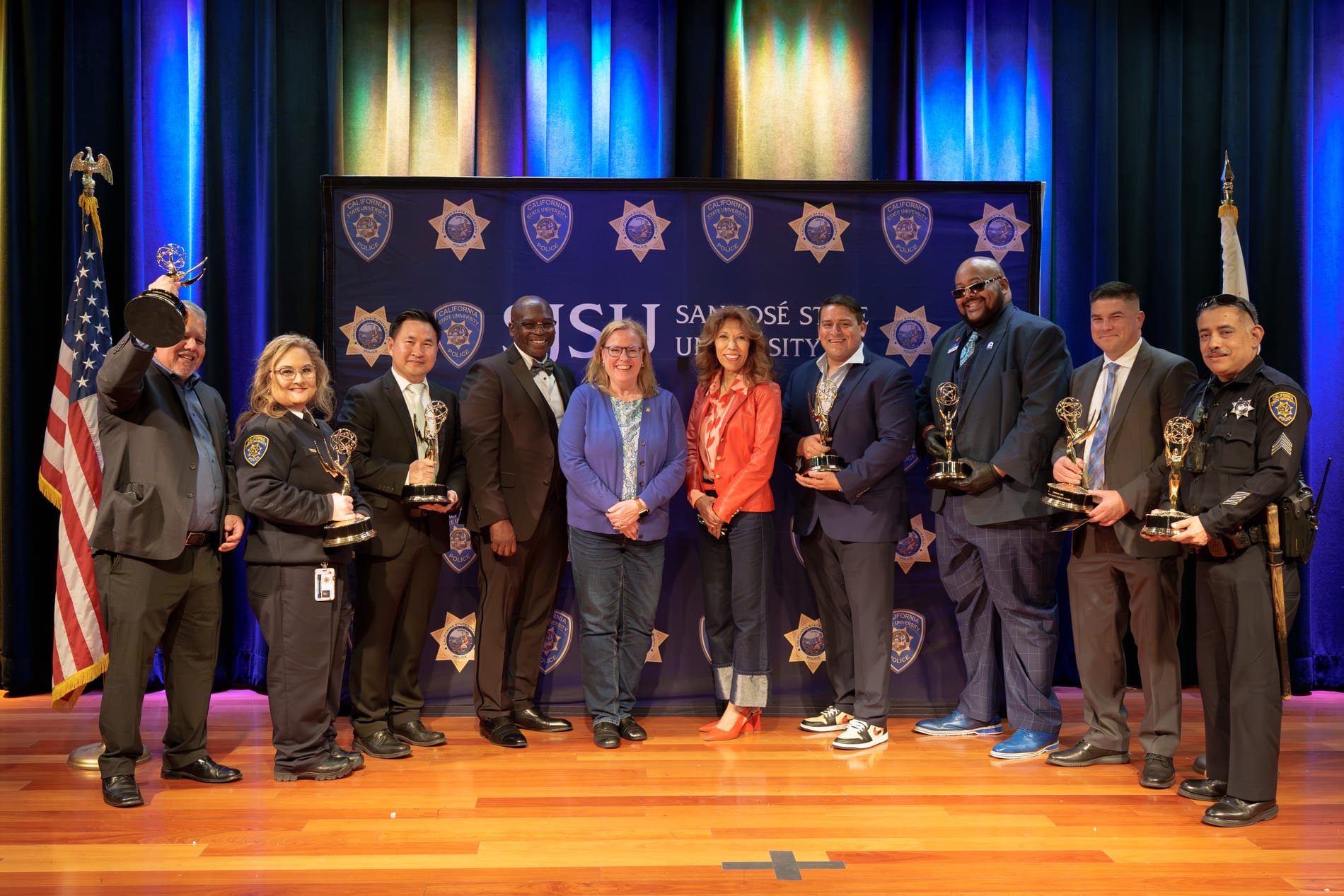 Ten award recipients and officers pose in a row on stage holding their Emmy statuettes, flanked by the American and California flags in front of a California State University Police backdrop