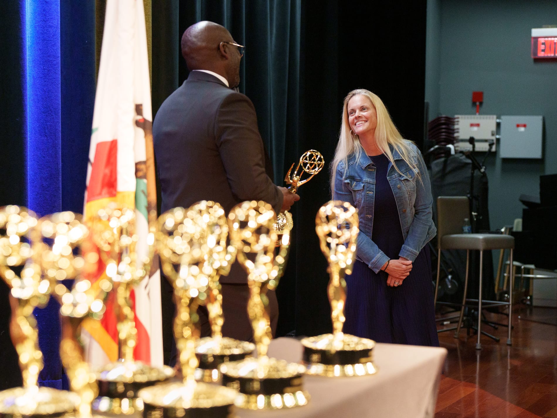 An officer chats with a guest in a denim jacket backstage, a table lined with Emmy statuettes blurred in the foreground