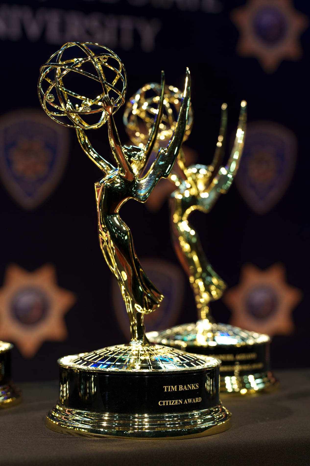 Two Emmy statuettes stand on a table, the nearest engraved Tim Banks Citizen Award, with a police badge backdrop blurred behind them