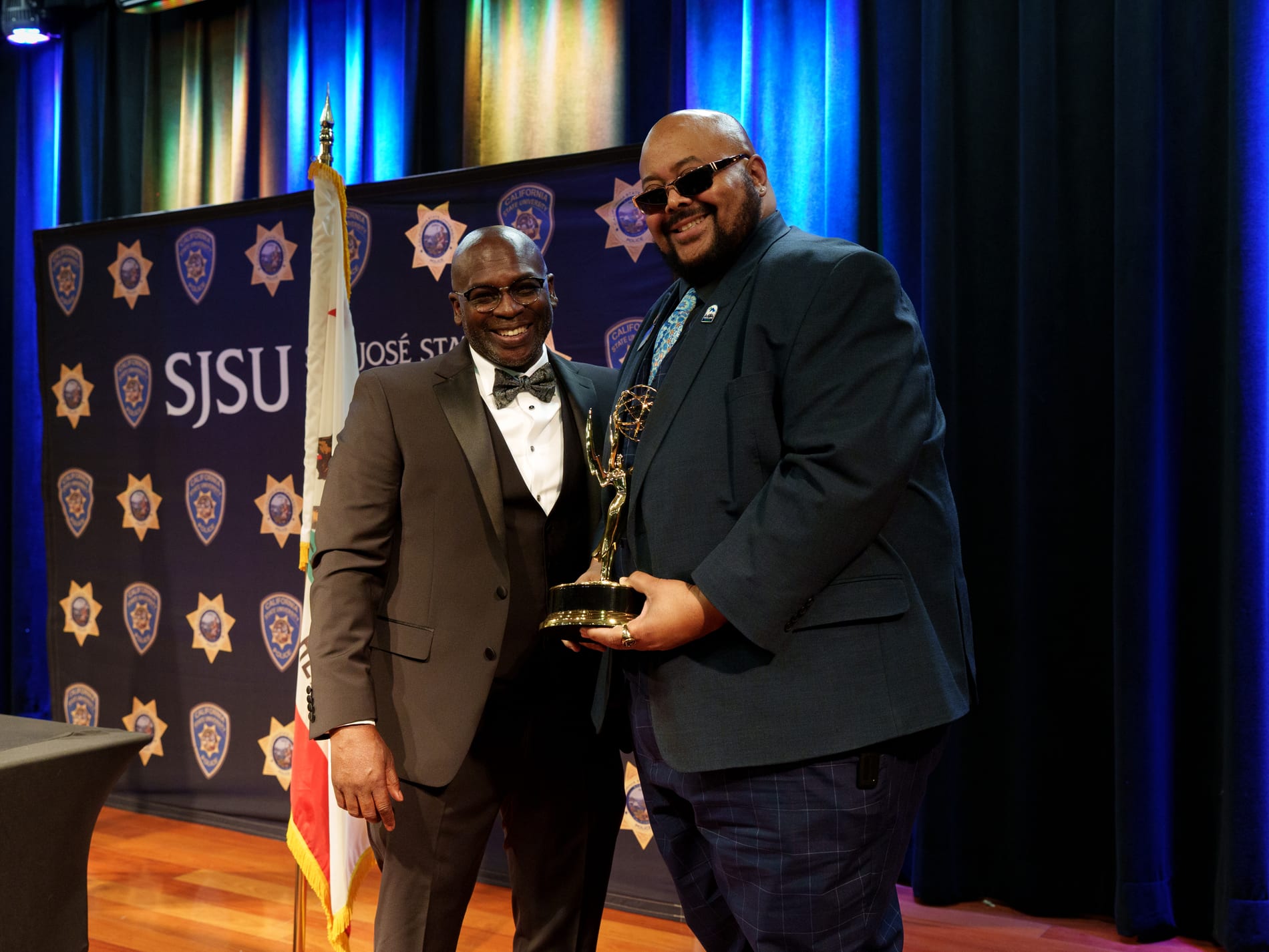 Two men pose together on stage smiling, one in a tuxedo bow tie and the other holding an Emmy statuette, in front of the SJSU backdrop