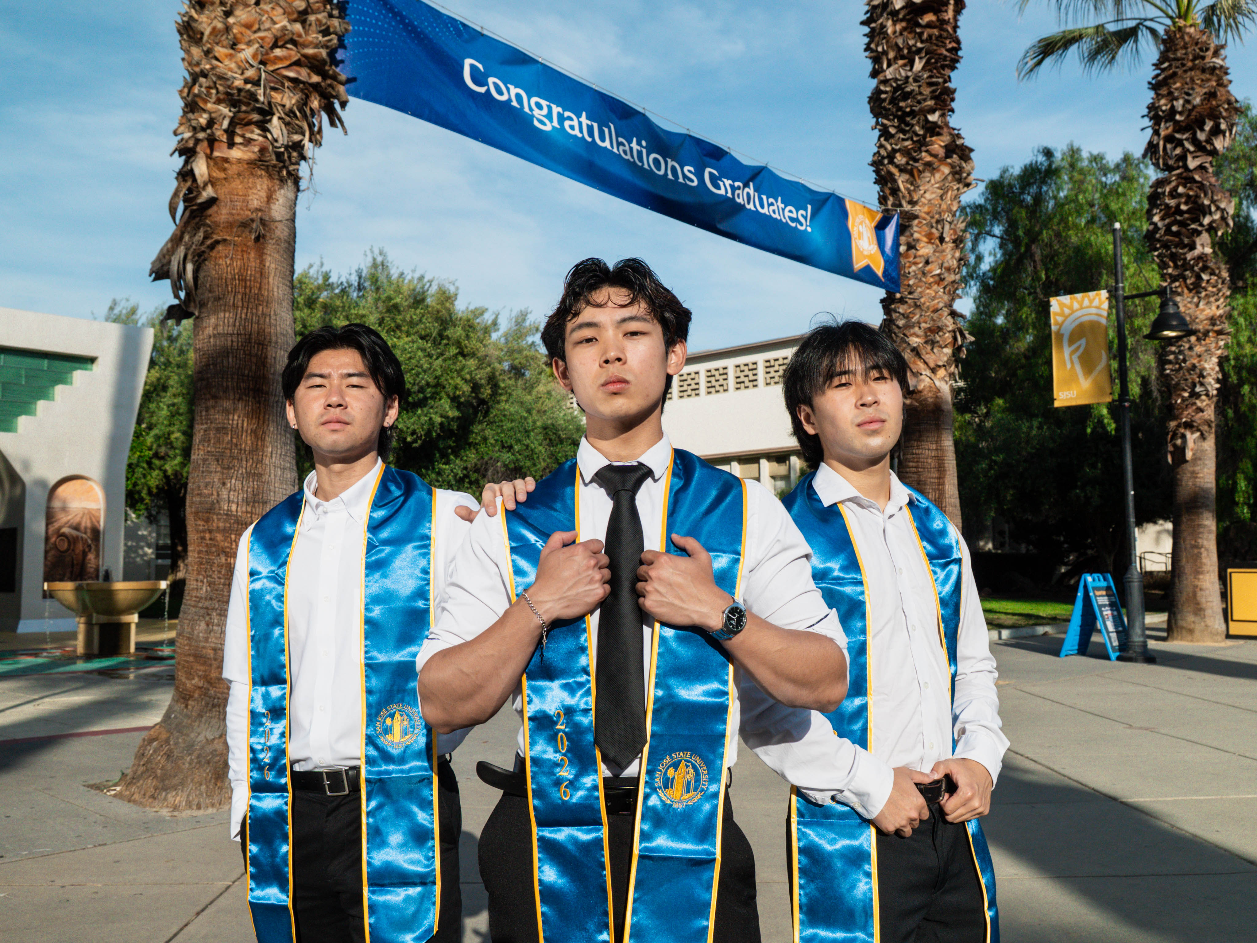 Three San Jose State graduates in blue and gold stoles under a Congratulations Graduates banner
