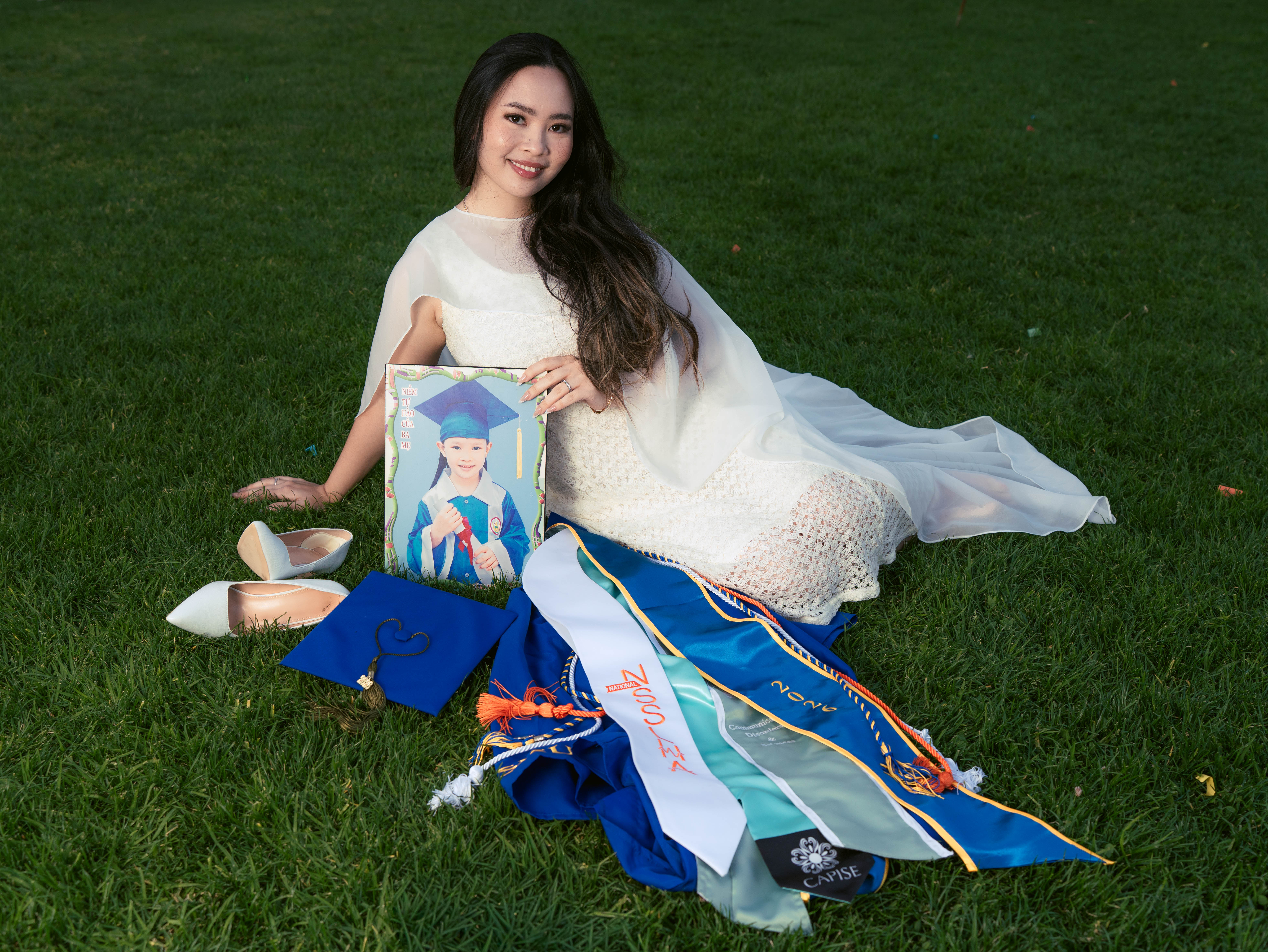 SJSU graduate lying in the grass with her cap, gown, and honor cords laid out beside her