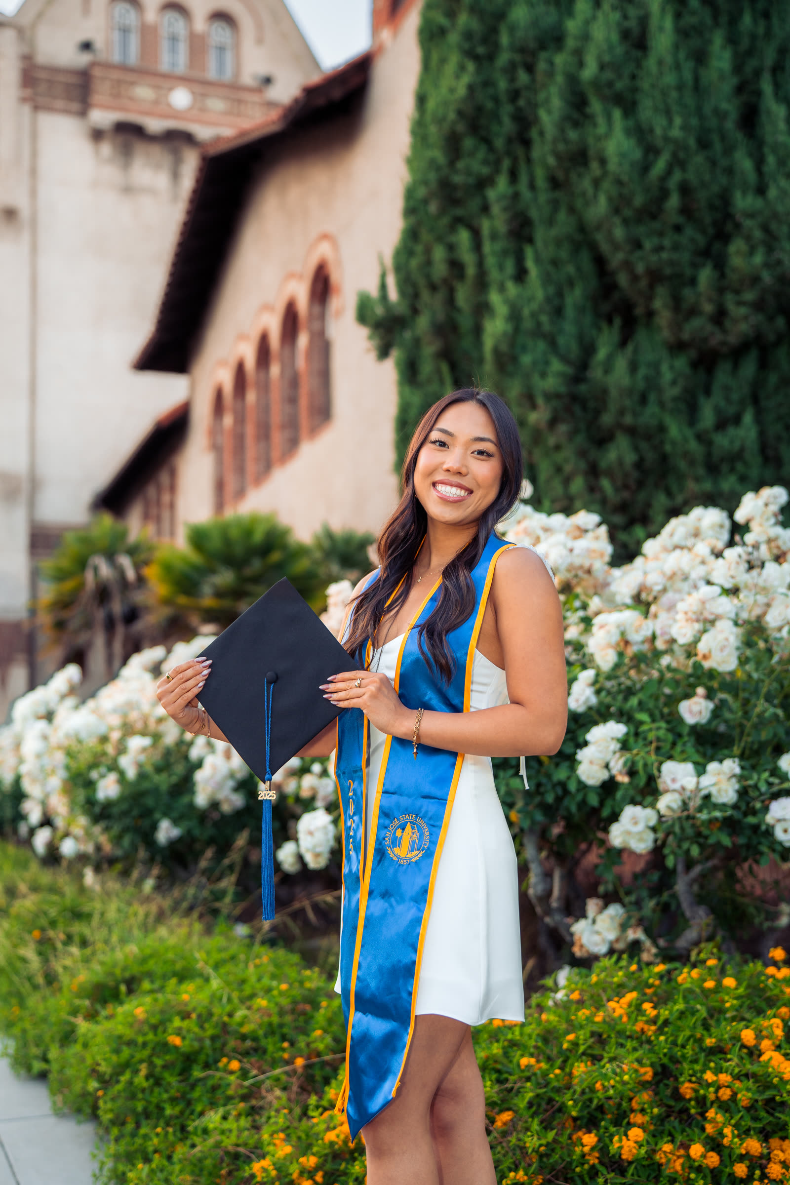 SJSU graduate holding her cap in front of blooming roses on campus