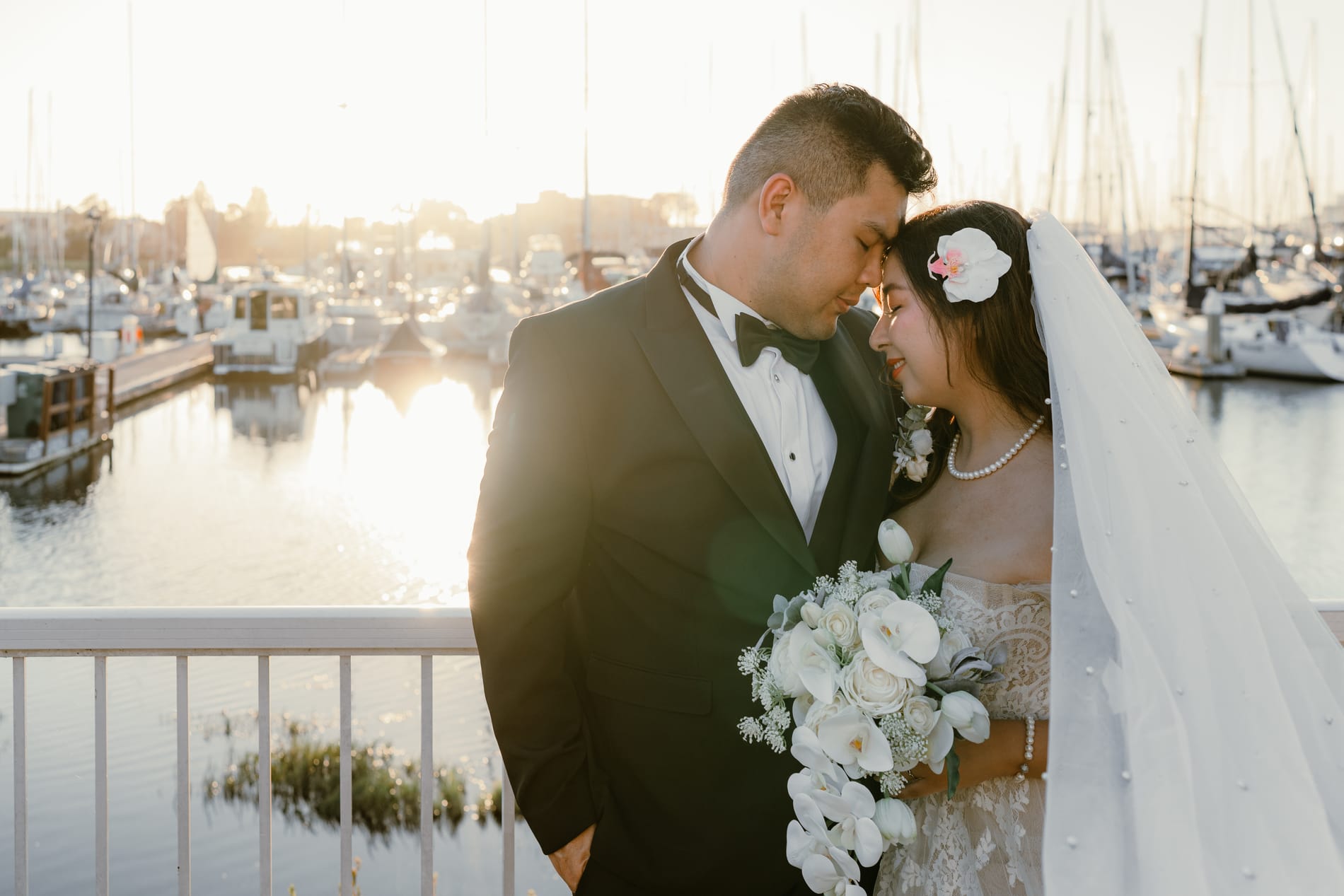 Bride and groom leaning into each other with her white bouquet between them, sun flaring off the marina
