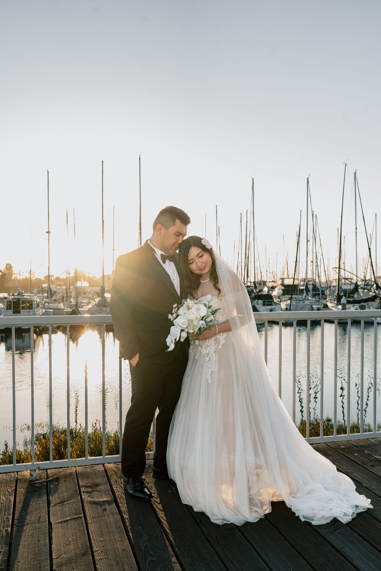 Bride resting against the groom on the dock, sailboat masts and still water behind them