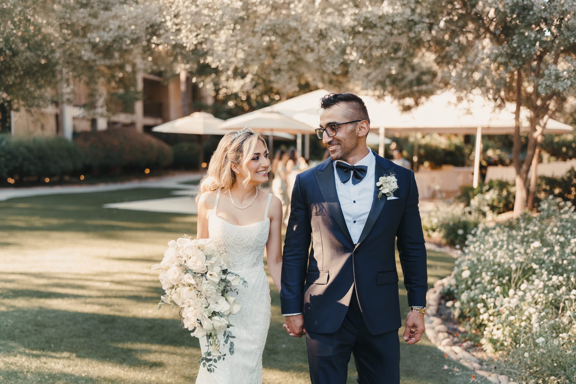 Bride and groom walking hand in hand across the lawn beneath the olive trees