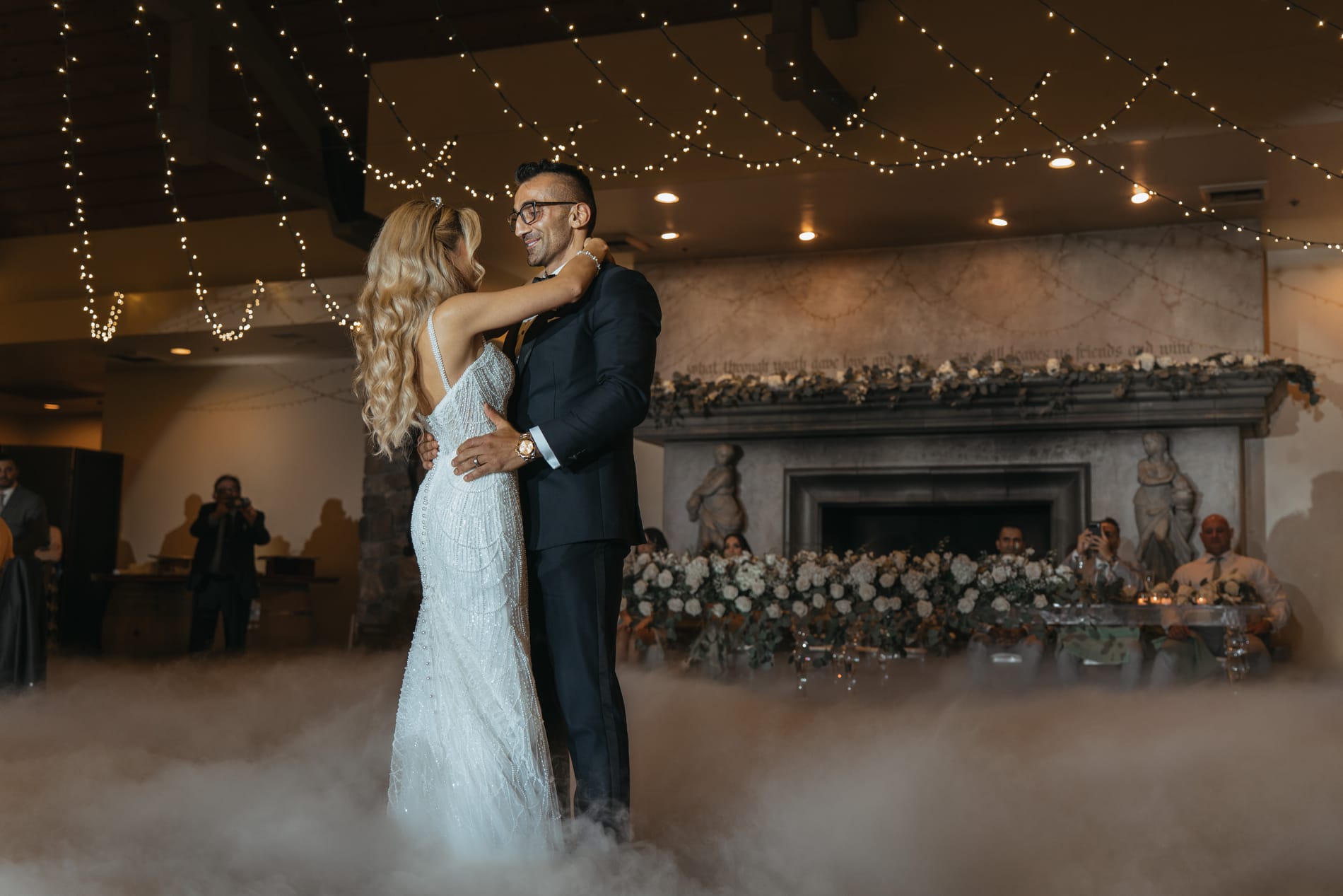 First dance in low fog beneath string lights, guests watching from the fireplace