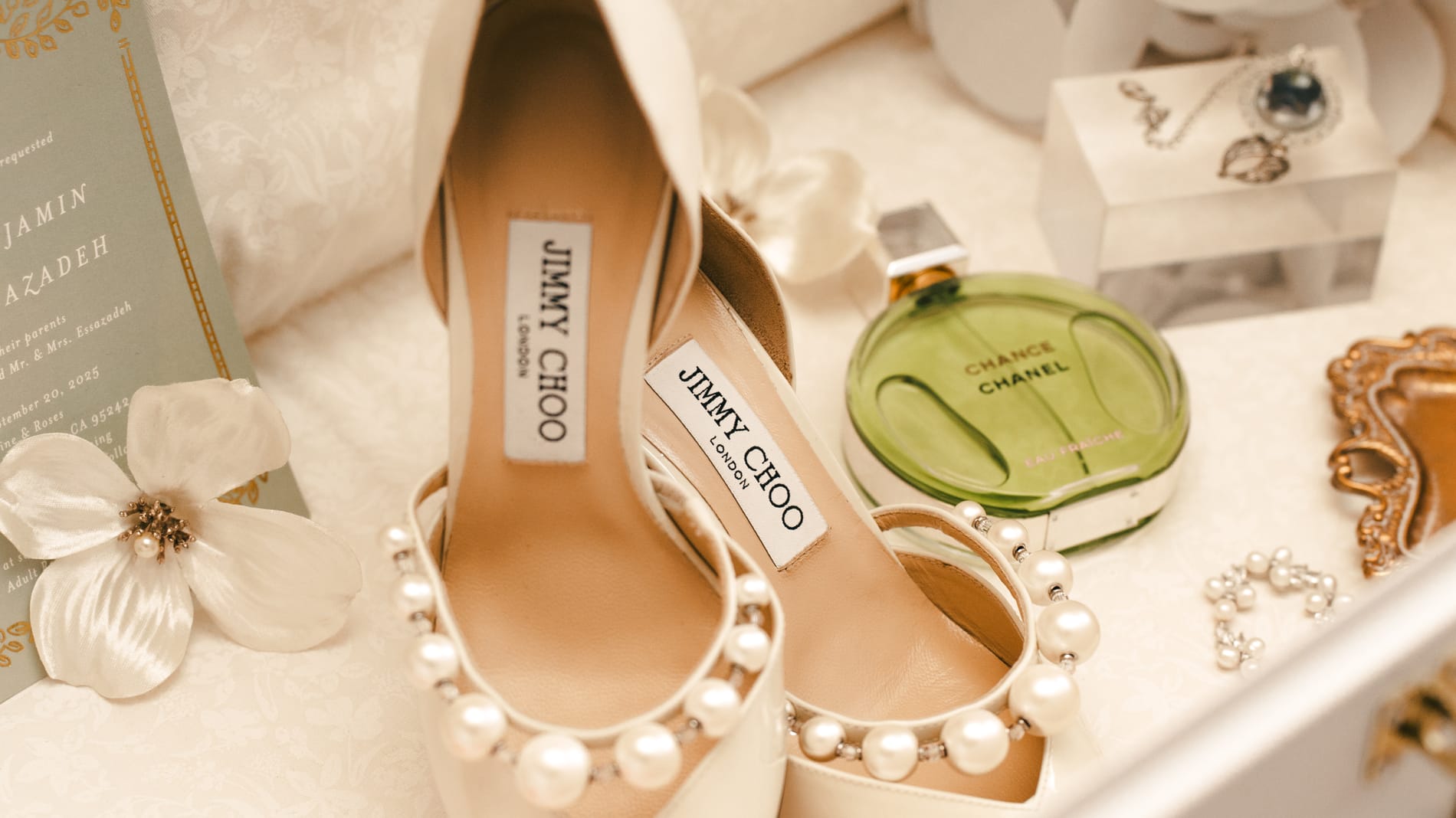 Bridal details flat lay: pearl-strapped Jimmy Choo heels, Chanel perfume, jewelry and the invitation