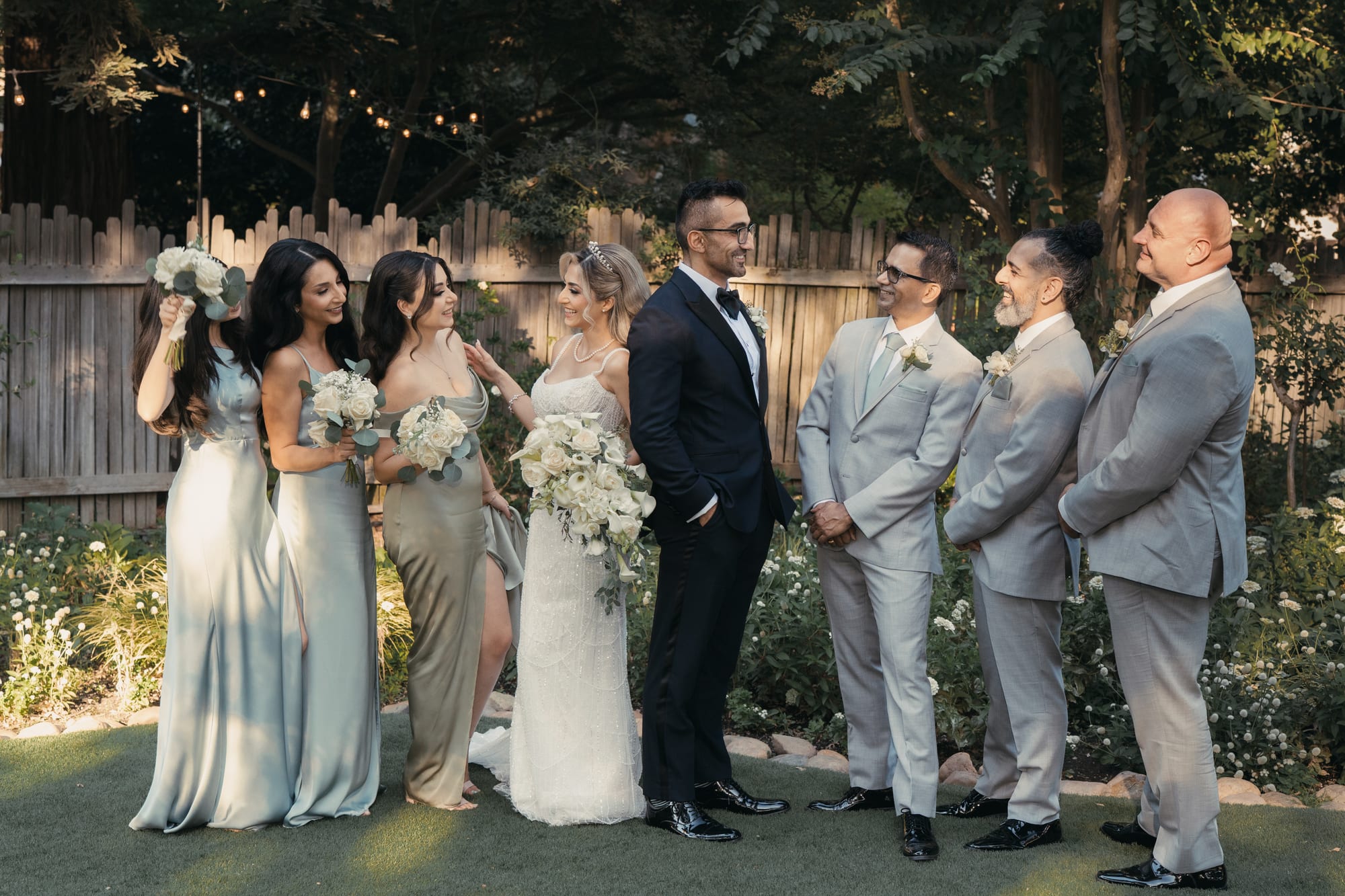 Bride and groom laughing with their wedding party in the garden, bridesmaids in sage satin and groomsmen in gray