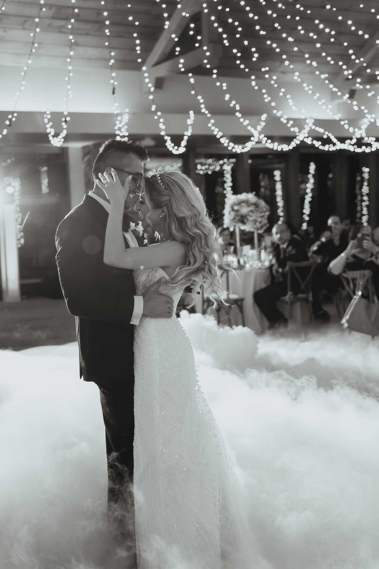 First dance beneath string lights, fog across the floor, black and white